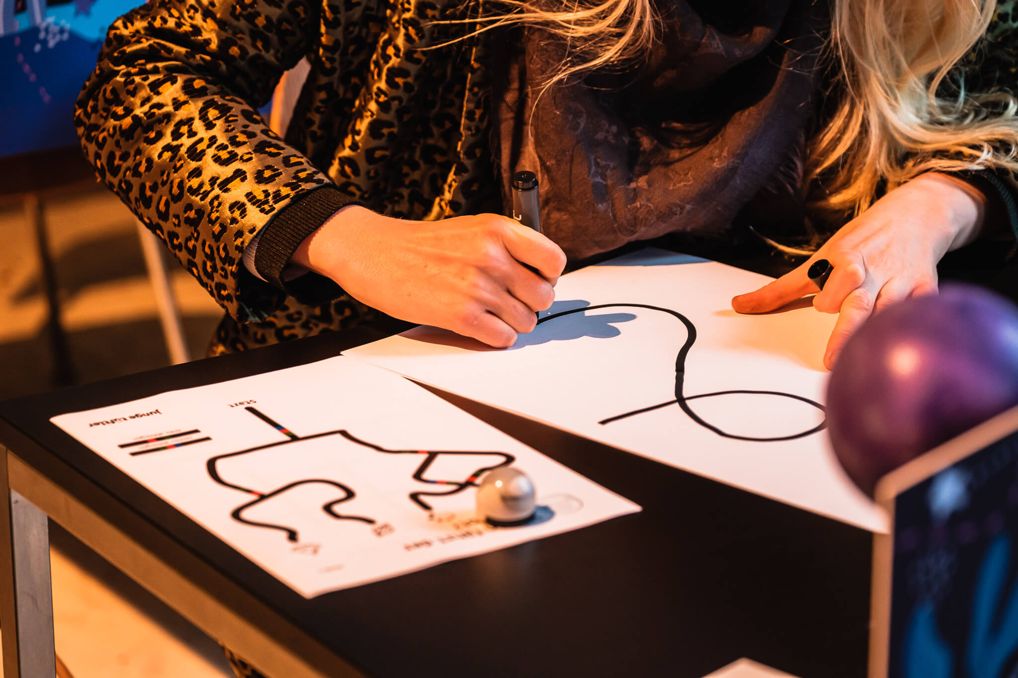 Kind malt eine schwarze Linie auf ein Blatt Papier, daneben liegt ein Ozobot-Arbeitsblatt mit einem Ozobot