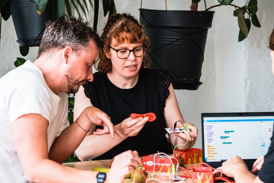 Zwei Personen experimentieren mit einem Makey Makey und Kabeln an einem Tisch, daneben ein Laptop der Programmiercode anzeigt