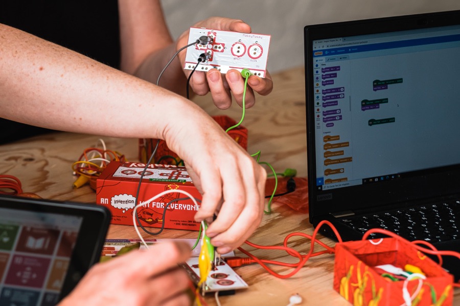 Zwei Hände halten ein Makey-Makey-Board mit Kabeln und Krokodilklemmen.
