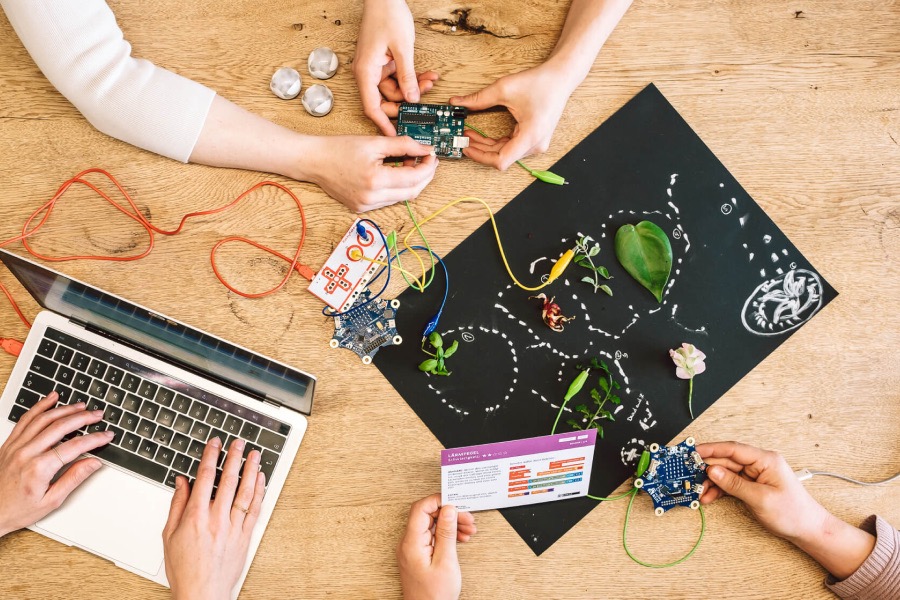 Mehrere Personen experimentieren gemeinsam an einem Tisch mit Laptop, Mikrocontrollern, Kabeln und Naturmaterialien.