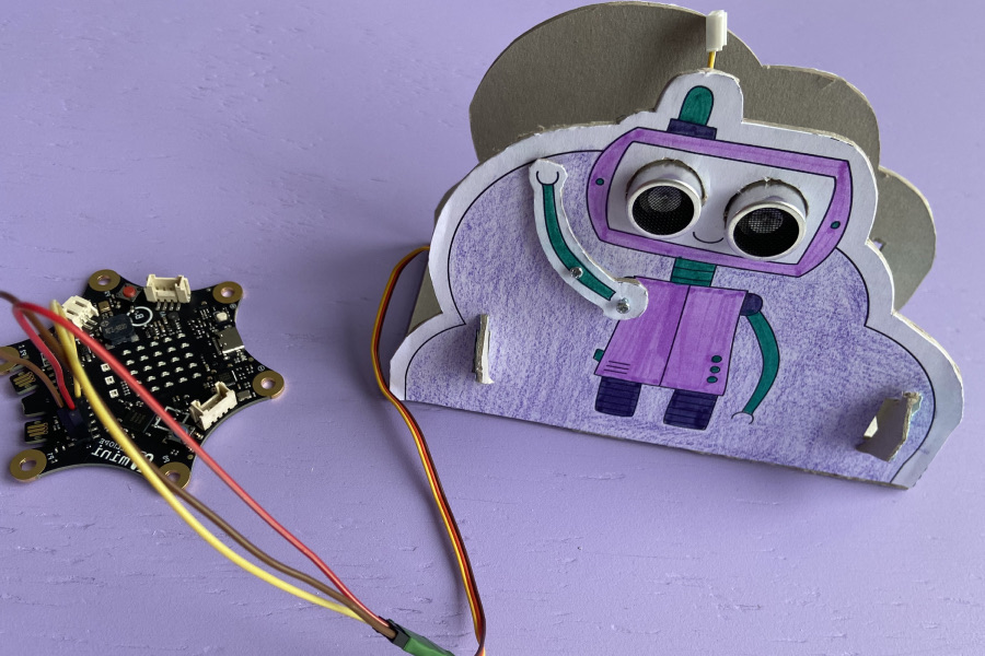 Ein aus Pappe gebastelter Roboter, sein Arm ist beweglich. Der Roboter ist mit einem Calliope mini durch zwei Kabel verbunden