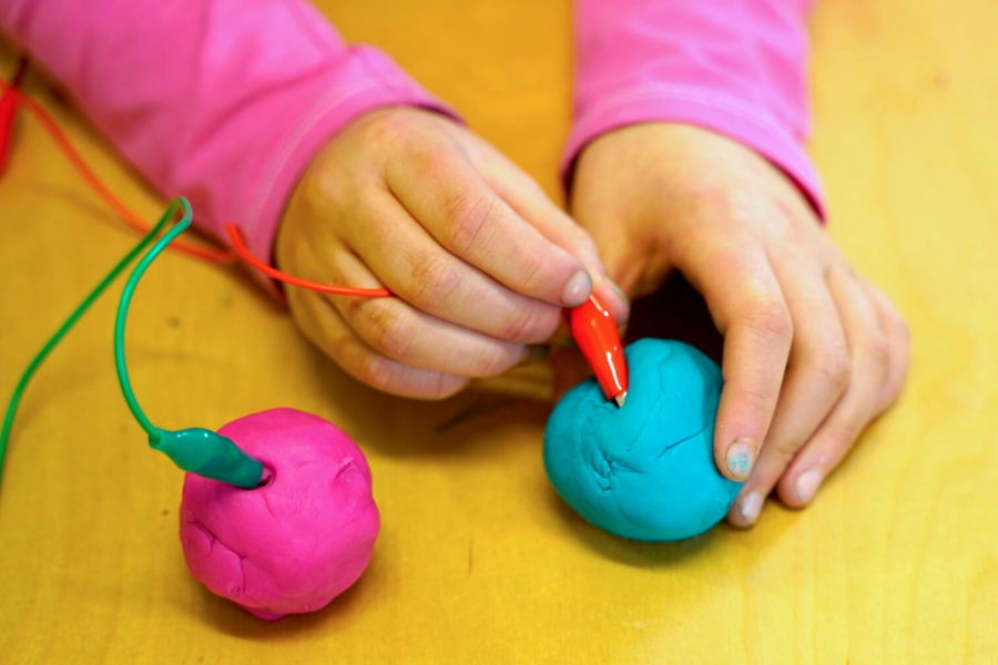 Kind steckt ein Verbindungskabel in einen Ball Knete, daneben liegt ein zweiter Ball Knete mit einem Verbindungskabel drin.